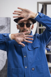 Porträtt på konstnären JR iklädd en blå skjorta, solglasögon och hatt. Händerna är placerade i luften framför ansiktet. 